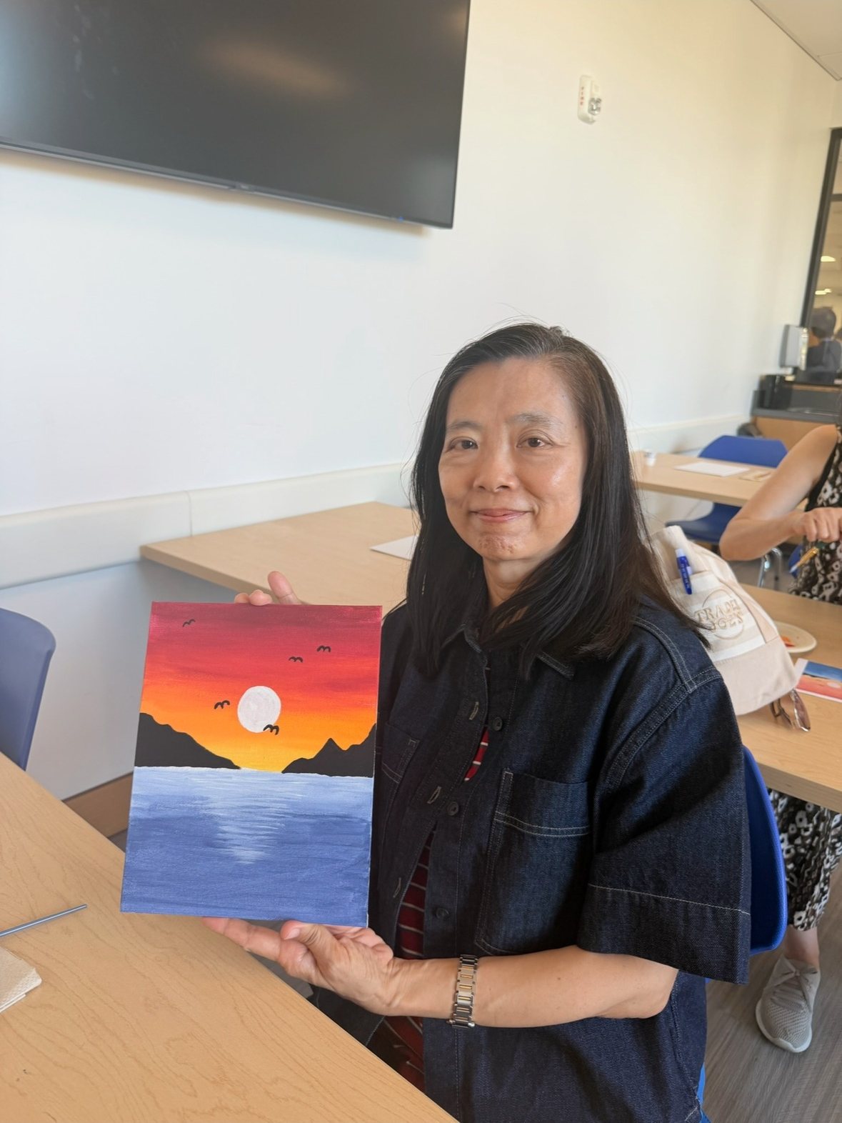 A participant smiling and holding up her sunset painting of orange and pink skies over a blue sea