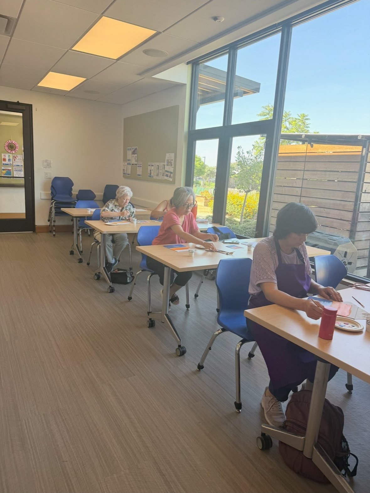 A bright workshop room with participants painting at tables by large windows