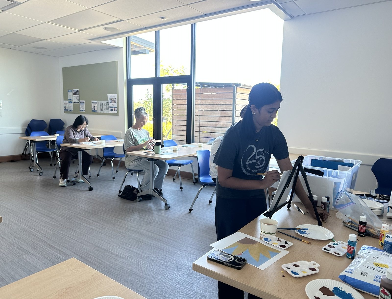 Participants painting at tables in a sunny room while a CARES officer sets up an easel