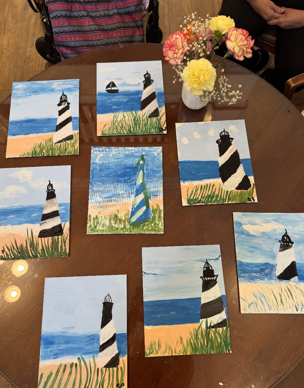 A round table covered with finished lighthouse paintings and a vase of flowers
