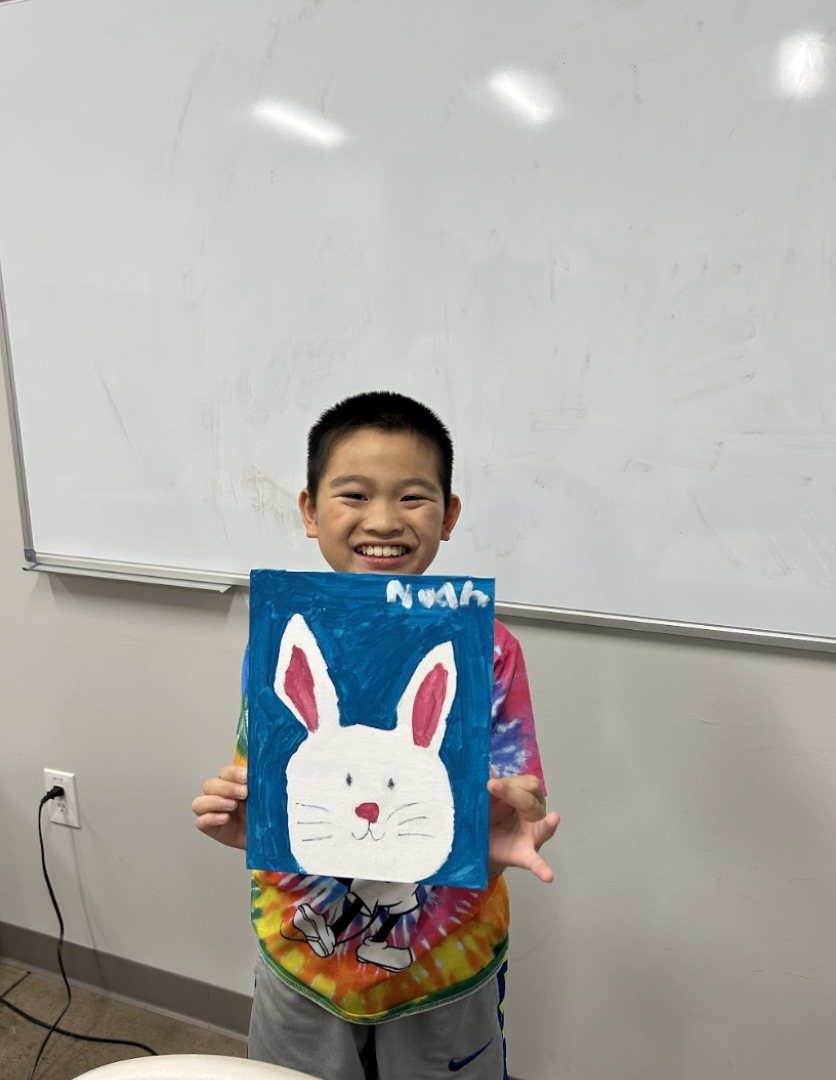 A participant smiling and holding up his white bunny painting on a bright blue background