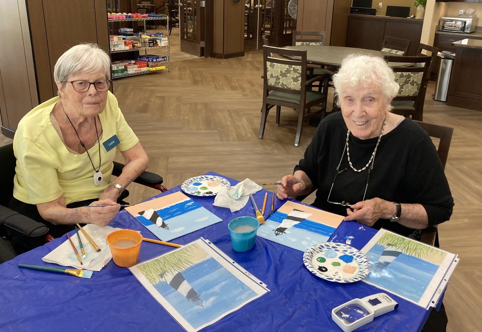 Two residents smiling and painting lighthouse scenes at a table with a blue tablecloth