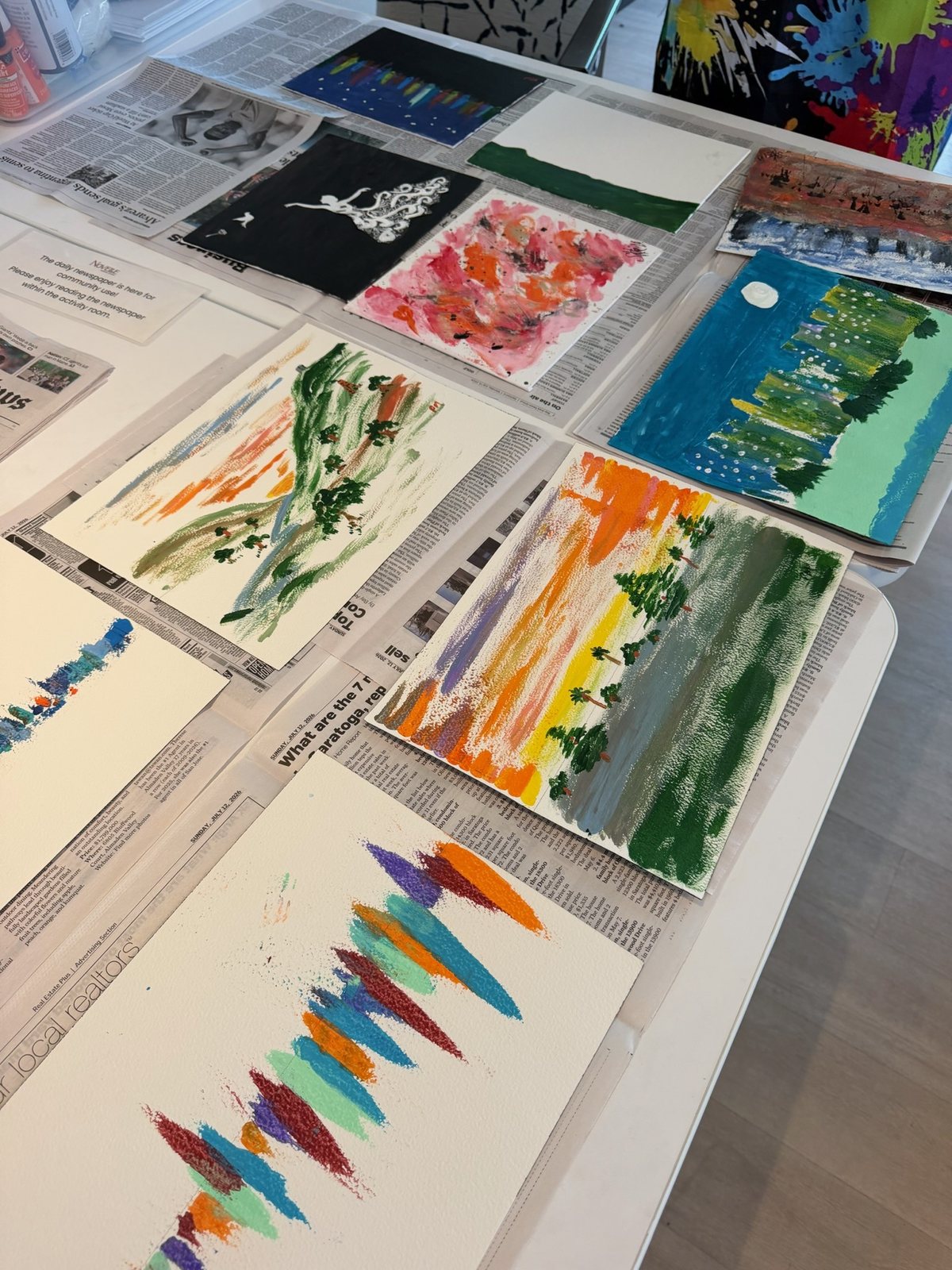 A table covered with colorful, expressive paintings drying on newspaper